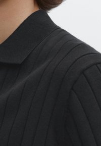 Musta ribattava polo-paita, jossa on kauluksenomainen kaulus ja tekstuuri, korostaen pystysuoria linjoja istuvassa designissa.