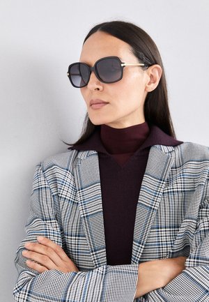 Fekete, oversized napszemüveg szürke lencsékkel, arany kiegészítőkkel és téglalap alakban. Kockás blézert és sötét garbót visel.