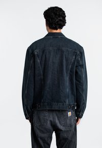 Giubbotto in denim di colore scuro con una vestibilità rilassata. Caratterizzato da cuciture visibili, due tasche laterali e bottoni in metallo. La vista posteriore mette in mostra un design classico.