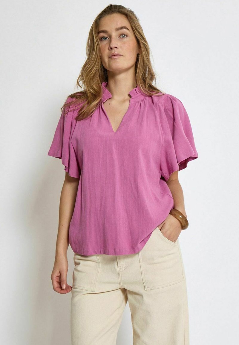 Blusa rosa a maniche corte con scollo a V e spalle plissettate, realizzata in tessuto leggero. Abbinata a pantaloni a gamba larga color beige chiaro.
