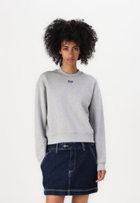 Gray sweatshirt with a round neckline and ribbed cuffs, featuring a blue logo patch. Paired with a dark denim skirt with pockets.