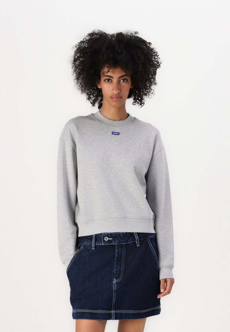 Gray sweatshirt with a round neckline and ribbed cuffs, featuring a blue logo patch. Paired with a dark denim skirt with pockets.