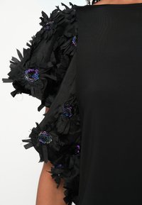 Musta paita, jossa on teksturoitu kukkakoriste hihassa. Hihassa on röyhelömäistä kangasta ja sinisen sekä violetin sävyisiä paljettikoristeita.