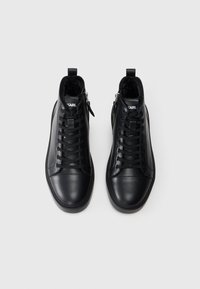 Svarte høyhælte sneakers laget av glatt lær, med snøring foran, avrundet tå og glidelås på siden. Fôret med mykt svart materiale.