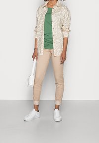 Béžová košile s vzorem, zelené tričko, béžové tapered kalhoty a bílé tenisky. Nese malou bílou tašku. Zobrazena volnočasová móda.