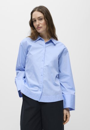Femme aux longs cheveux bruns portant une chemise ample bleu clair à boutons et un pantalon noir, debout avec une main dans la poche.