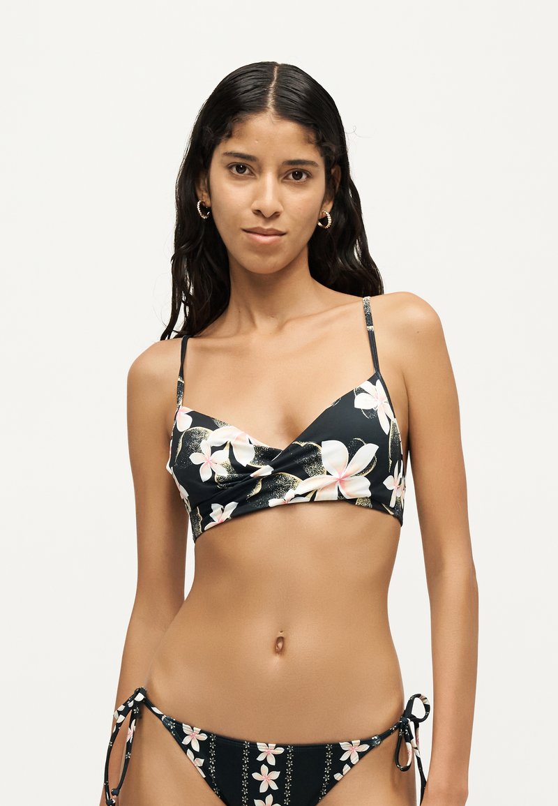 Femme aux longs cheveux foncés portant un bikini noir à fleurs blanches et roses, debout devant un fond uni clair.