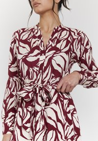 Maroon- und cremefarbenes Hemdkleid mit botanischem Druck, Taillenzug, langen Ärmeln und kragenlosem Ausschnitt. Weicher Stoff mit glatter Textur.