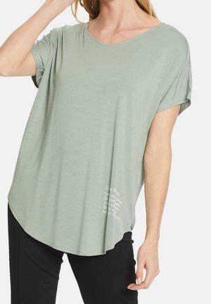 Frau trägt ein lockeres, kurzärmeliges, hellgrünes Oberteil mit kleinem weißen Text nahe dem Saum, kombiniert mit schwarzen Hosen, vor neutralem Hintergrund.