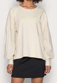 Felpa oversize beige chiaro con maniche lunghe a palloncino. Presenta una tasca frontale e un tessuto testurizzato. Abbinata a una gonna nera.