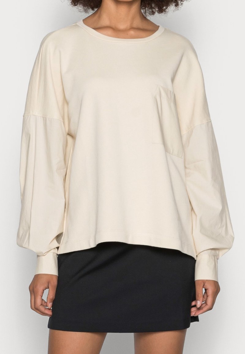 Felpa oversize beige chiaro con maniche lunghe a palloncino. Presenta una tasca frontale e un tessuto testurizzato. Abbinata a una gonna nera.