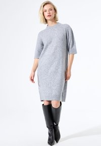 Femme aux cheveux blonds portant une robe en laine grise longueur genoux avec des manches 3/4 et des bottes noires à talons hauts, se tenant debout.