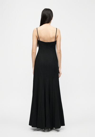 Femme aux longs cheveux noirs portant une robe noire longue jusqu'au sol avec de fines bretelles, vue de dos sur un fond uni.