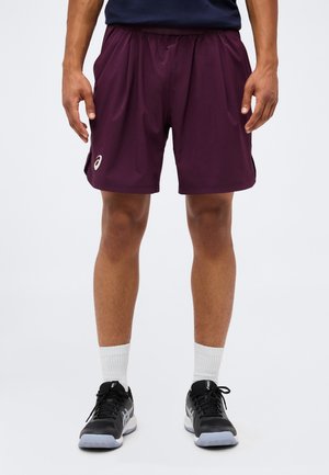 Homme portant un short de sport violet, des chaussettes blanches montantes et des chaussures de course noires, debout devant un fond uni.