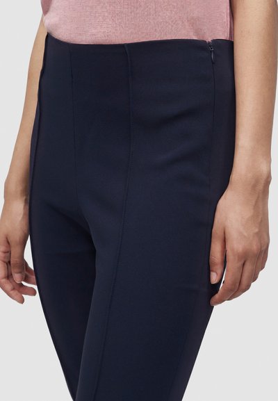 Gros plan d'une personne portant un pantalon bleu marine ajusté avec une fermeture éclair latérale et un haut rose, les mains détendues sur les côtés.