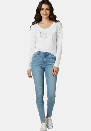 Giovane donna che indossa una maglietta bianca a maniche lunghe con scollo a V e logo, jeans skinny azzurri chiaro e sneakers bianche, in piedi davanti a uno sfondo semplice.