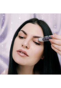 Bottiglia a sfera in vetro trasparente con cristalli viola all'interno, applicata sulla pelle. Il modello ha lunghi capelli scuri e una pelle liscia. Sfondo morbido.