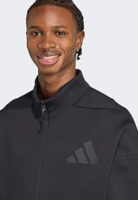 Chaqueta negra de cremallera con cuello alto, que cuenta con un acento de logo mate y una tela lisa y texturizada.