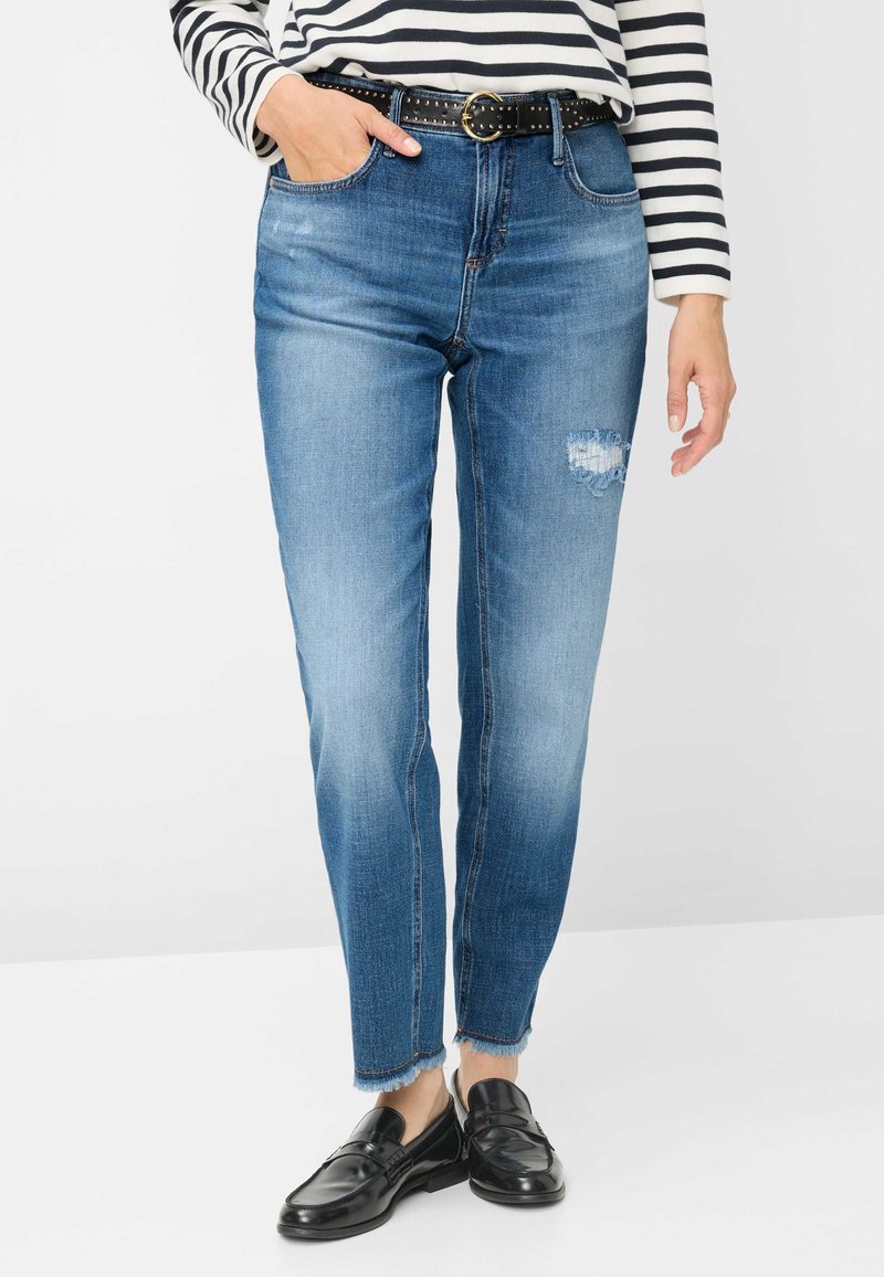 Jeans di denim blu con finitura sbiadita, caratterizzati da uno strappo sulla coscia destra, taglio dritto e orlo sfrangiato, abbinati a una cintura nera e mocassini.