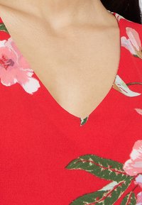 Top vermelho de decote em V com um padrão floral, apresentando flores cor-de-rosa e folhas verdes; textura suave e design casual, com ênfase no decote e na estampa.