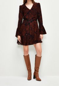 Femme portant une robe marron à manches longues avec des poignets évasés et une fine ceinture noire, associée à des bottes marron à talons montant jusqu'au genou.