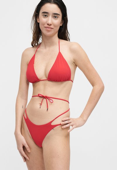 Γυναίκα που στέκεται και φορά κόκκινο μικροσκοπικό μαγιό με κορδόνια πάνω και κάτω, πάνω σε απλό ανοιχτό φόντο.