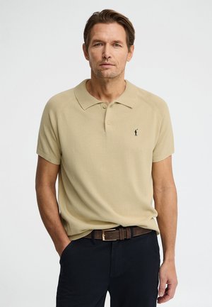 Man draagt beige polo met korte mouwen en kraag, donkere broek en bruine riem, en staat met één hand in de zak tegen een witte achtergrond.