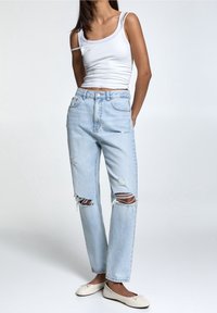 Donna che indossa un top senza maniche bianco, jeans strappati blu chiaro e ballerine bianche, in piedi con le mani dietro la schiena su uno sfondo neutro.