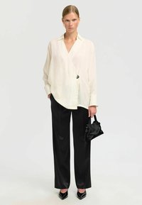 Blouse blanche et légère de style wrap avec un seul bouton noir, associée à un pantalon noir taille haute et un petit sac à main noir.