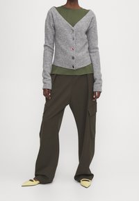 Šedý vlněný cardigan s výstřihem do V, zapínání na pět knoflíků, navlečený přes zelený top. Volné olivově zelené kalhoty a žluté špičaté boty.