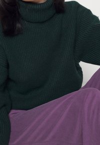 Maglione a collo alto in maglia verde scuro con texture a costine, abbinato a una gonna fluente viola. Focalizzati sui dettagli del tessuto, sul colore e sul motivo.
