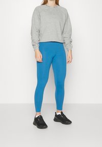 Felpa grigia con maniche raglan, abbinata a leggings blu. Le sneakers nere completano il look. Trame morbide e design semplice.