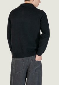 Maglione nero lavorato a maglia con colletto e polsini a coste, indossato su pantaloni di jeans grigi con due tasche posteriori. Design semplice e funzionale.