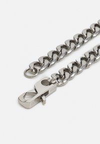 Accessoire en chaîne métallique avec des maillons entrelacés. Présente un fermoir robuste et une finition en argent poli. Les maillons sont lisses et plats.