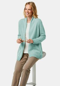 Helles mintgrünes Cardigan mit Schalkragen, gerippten Bündchen und zwei vorderen Taschen, getragen über einem weißen Oberteil und kombiniert mit beigen Hosen.