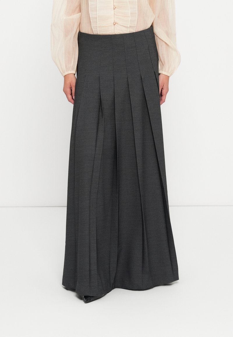 Femme portant un pantalon plissé à jambes larges gris foncé et un chemisier beige clair à manches longues avec des détails froncés sur le devant.