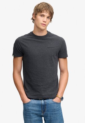 Jonge man met kort lichtbruin haar, die een donkergrijs T-shirt, blauwe spijkerbroek en een polshorloge met een bruine leren band draagt.