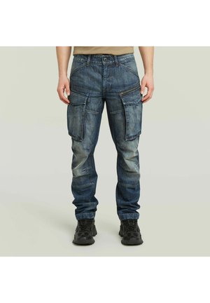 Uomo che indossa pantaloni cargo in denim blu sbiadito con più tasche e scarpe da ginnastica nere, in piedi contro uno sfondo chiaro e uniforme.