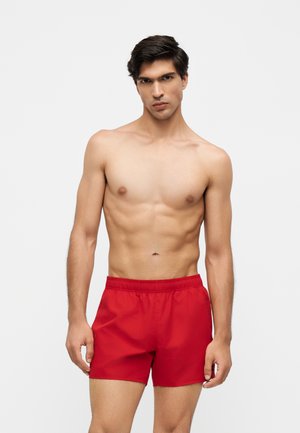 Jeune homme aux cheveux foncés portant un short de bain rouge vif, debout devant un fond clair uni, regardant devant avec une expression neutre.