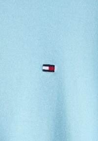 Jasnoniebieska bawełniana koszulka z małym haftowanym logo z czarnymi, białymi i czerwonymi paskami na lewej piersi. Prosty design, miękka faktura.