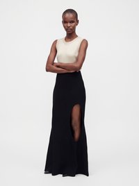 Woman wearing a sleeveless beige top and a long black skirt with a high slit, standing with arms crossed against a plain background.