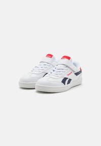 Zapatillas blancas con acentos en azul marino, que cuentan con una lengüeta acolchada y una correa de velcro, suela de goma texturizada y un logotipo rojo en la lengüeta.