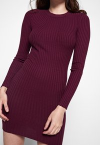 Donna che indossa un vestito a maniche lunghe, aderente e in maglia a coste di un profondo color prugna, in piedi contro uno sfondo bianco semplice.