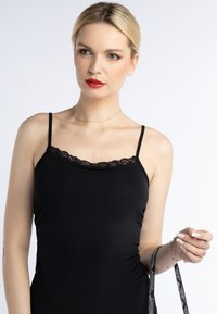 Camisola neagră cu dantelă la decolteu, bretele subțiri și o textură fină. Modelul ține un curea cu un model în una dintre mâini.