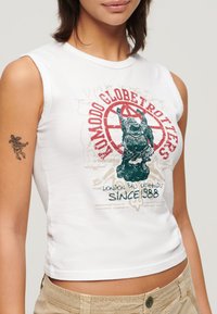 Top corto sin mangas de color blanco que presenta un estampado gráfico de una estatua, con acentos en rojo y verde, y el texto que dice "Londres Bali Katmandú Desde 1988."