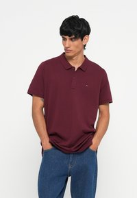 Burgunderfarget polo-skjorte med krage, to-knaps placket og liten logo-detalj. Båret med blå denim jeans. Korte ermer.