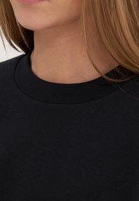 Close-up af en person iført en sort rundhalset t-shirt, der viser nakken, den nederste del af ansigtet og lyst brunt hår.
