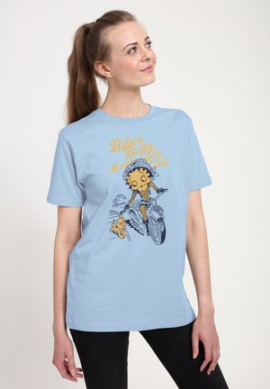 Kobieta w jasnoniebieskiej koszulce z vintage'ową ilustracją motocyklistki i psa oraz napisem "Biker Betty Motorcycle Club".