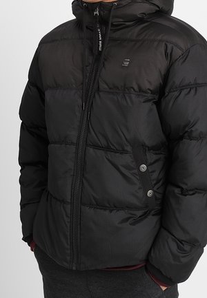 Doudoune noire avec sections matelassées horizontales, fermeture éclair à l'avant, poches à boutons-pression et capuche, portée par une personne les mains dans les poches.