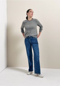 Harma neulepaita, jossa on pyöreä pääntie, yhdistetty sinisiin suoriin farkkuihin ja valkoisiin tenniskenkiin. Yksinkertainen muotoilu, rento istuvuus ja minimaalinen yksityiskohtaisuus.